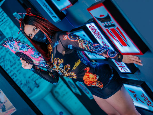 ValeryHarllow with red-brown hair, black mask, sheer black top with colorful embroidery, black shorts, holding pink and black fan, neon lights in background, looking at camera.