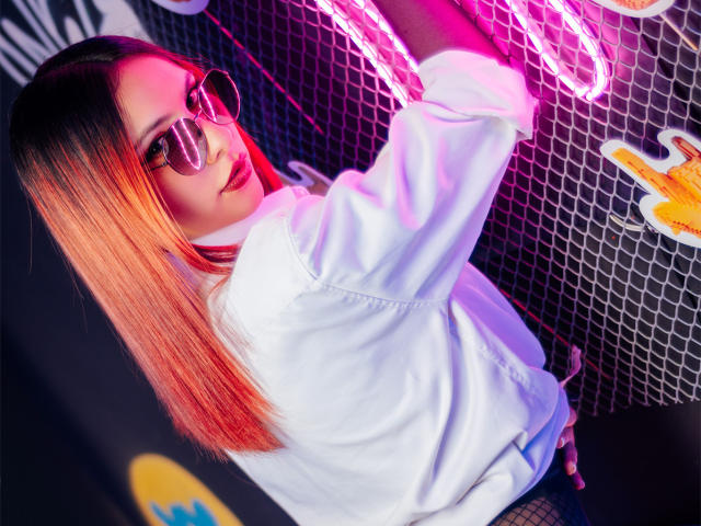 ValeryHarllow with long orange-brown hair, wearing a white hoodie, black fishnet pants, and reflective sunglasses, touching a neon pink sign. Pink and purple lighting highlights her.