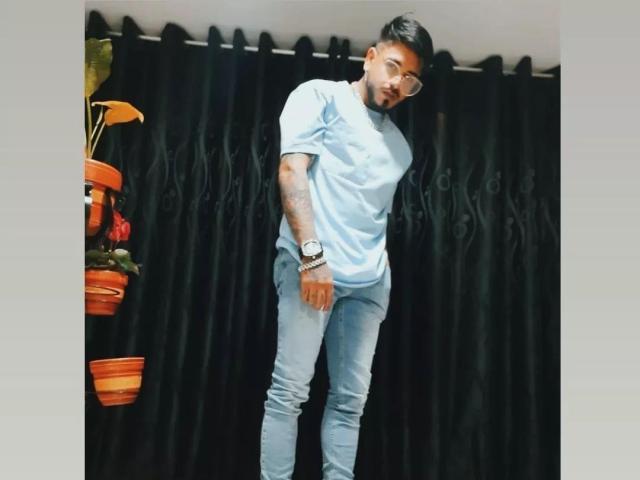 VisiI stands with a slight lean, wearing a light blue t-shirt and skinny jeans, tattoos on his right arm, and a watch on his left wrist.