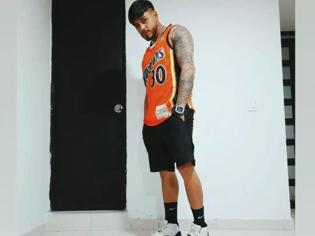 VisiI with tattoos on arms, wearing an orange "Los Angeles" basketball jersey, black shorts, socks, and white sneakers, standing in front of a black door.