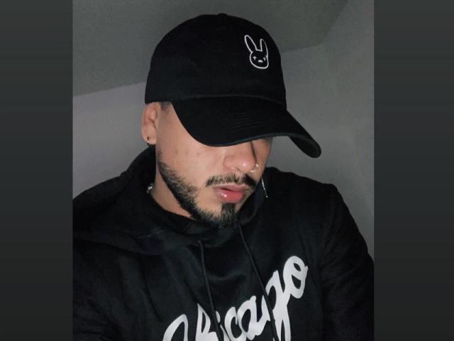 VisiI porte une casquette de baseball noire avec un logo de lapin blanc, une barbe, un anneau de nez et un sweat à capuche noir. Le visage de VisiI est partiellement caché par la casquette..