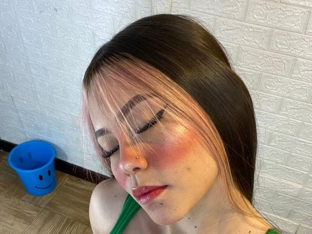 Jazmin Wells, con capelli rosa e neri, fard rosa, top verde, piercing al naso e un leggero alone di sporco sul viso, occhi chiusi, che guarda in basso, un secchio blu con una faccina sorridente sul pavimento..
