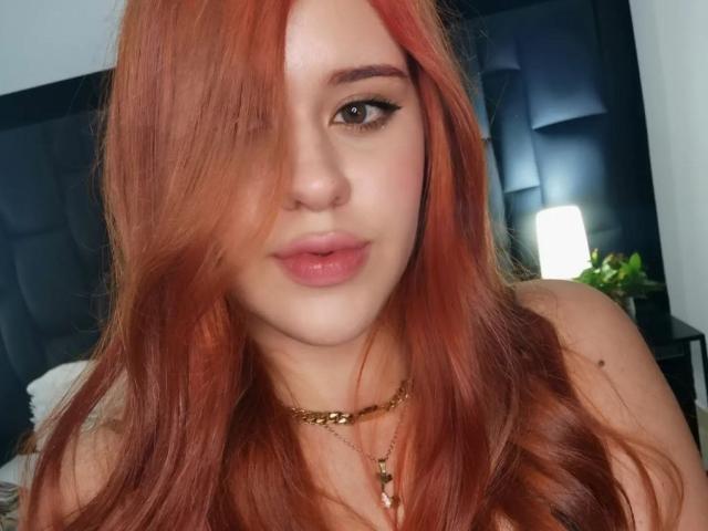AlessaClark with red hair, green eyes, and pink lips, wearing a gold necklace, has a slight pout, and her hair partially covers her right eye.