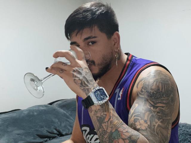 Une personne avec des tatouages, des cheveux foncés et des boucles d'oreilles, buvant dans un verre de vin, portant un maillot de basket violet et une montre, avec une expression sérieuse..