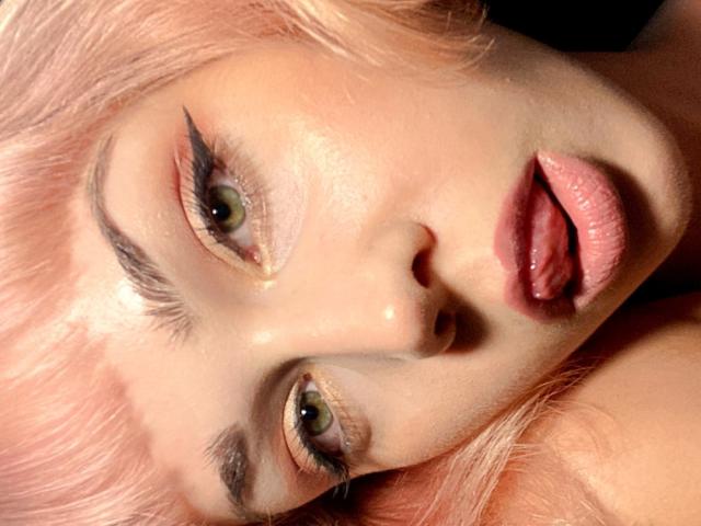 Le visage de LexiexWild, avec ses cheveux roses, ses yeux verts, son trait d'eye-liner prononcé et ses lèvres légèrement entrouvertes de couleur rose, avec la langue tirée. Sa peau est brillante, et elle regarde directement l'appareil photo..