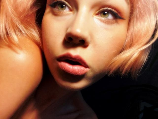 LexiexWild heeft roze haar, groene ogen en glanzende lippen met een licht geopende mond, en ze kijkt op met een sexy, verraste uitdrukking. Haar gladde, gebruinde huid wordt verlicht door zonlicht..