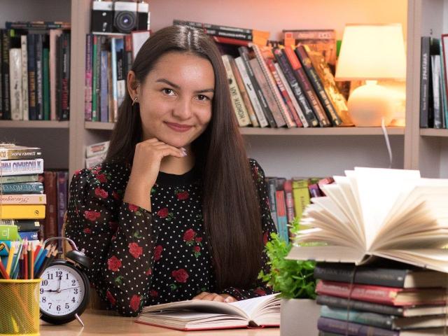 JasmineAsha, with long brown hair, wearing a black polka dot shirt with red roses, smiles at the camera, resting her chin on her hand, in a book-filled room.