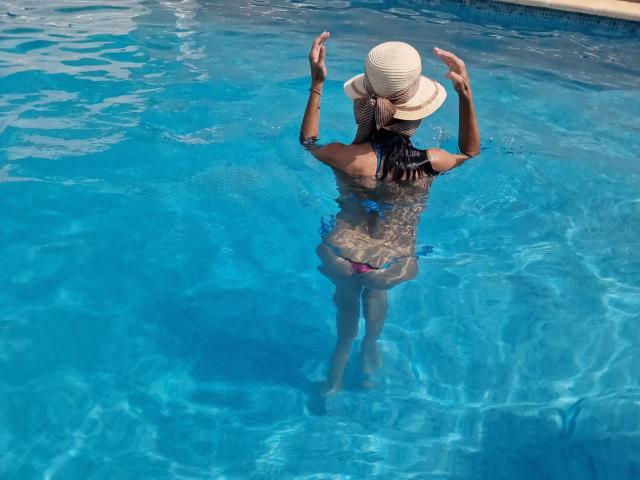 Cassandra Michelli se tient dans une piscine bleue, les bras levés, portant un chapeau de soleil et un maillot de bain rose, avec ses fesses et ses seins partiellement visibles à travers l'eau claire..