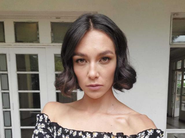 Belicious69 a les cheveux noirs et ondulés, coupés en un carré court, des yeux marron, et porte un haut noir à fleurs avec une épaule dénudée. Ses fesses ne sont pas visibles. Elle a l'air sérieuse..
