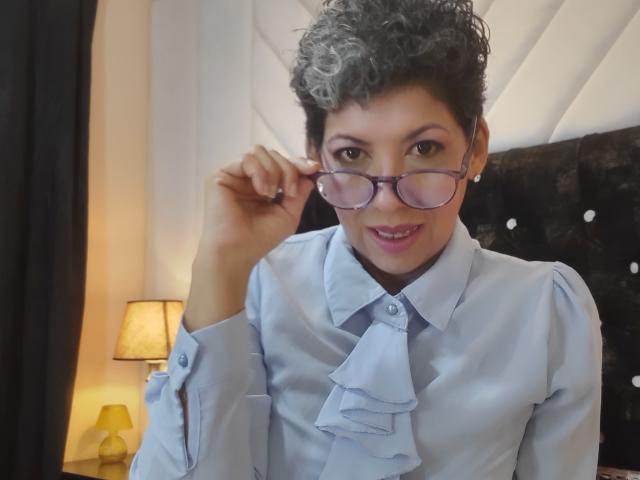 JuliaNewell with curly gray hair, wearing glasses, light blue shirt with ruffle, holding glasses, smiling, brown eyes, black headboard, yellow lamp, dark curtains, soft lighting.