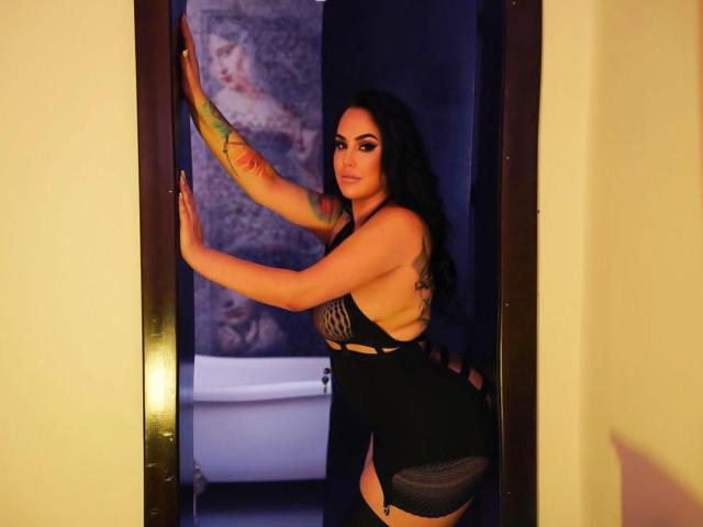 HottyNicole se tient dans un encadrement de porte, vêtue de lingerie noire, ses fesses et ses seins étant mis en valeur. Elle a des tatouages sur son bras et son côté, et ses longs cheveux noirs flottent..
