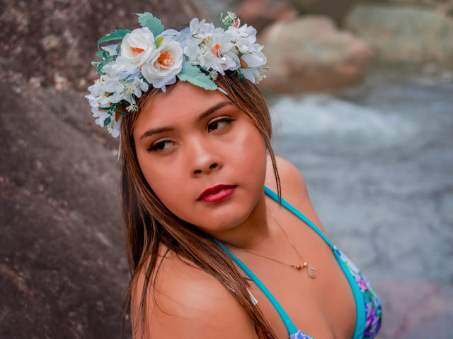 Eva Madison met bruin haar, een bloemenhoofddeksel, rode lippenstift, een blauw haltertopje, een ketting en een bruine huid, kijkt naar de camera, in de buurt van rotsachtig water. Borsten zijn gedeeltelijk zichtbaar, het water is onscherp op de achtergrond..