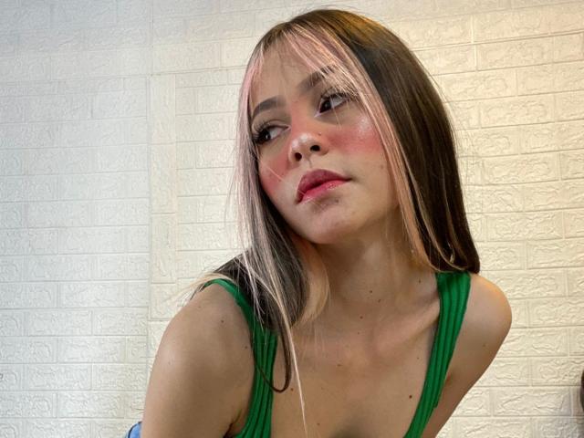 JazminWells heeft roze en bruin haar, een groene top, rode blush en een schattig gezicht met kleine borsten, ze kijkt iets naar beneden met een zachte glimlach. Haar huid is licht..