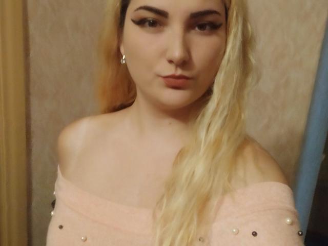 KrystaXlMagic ha i capelli biondi, l'eyeliner nero e un piercing al naso. Indossa una maglietta rosa con una spalla scoperta e piccole perle. Il suo seno è leggermente visibile. Sta guardando la fotocamera..