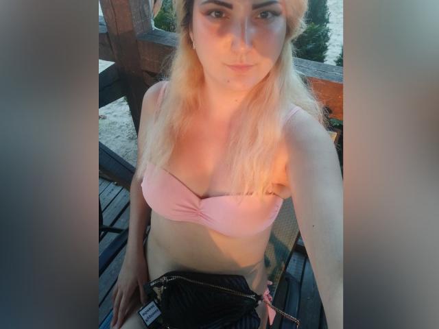 KrystaXlMagic com cabelo loiro, sutiã rosa, mostrando os seios, saia preta, em pé em um deck de madeira, olhando para a câmera, braços ao lado, luz do início da noite, sorriso leve..