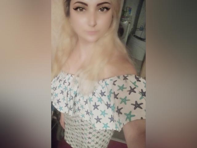 KrystaXlMagic con i capelli biondi, top a stella con spalline scoperte, seno piccolo, trucco leggero, che guarda la fotocamera, leggero sorriso, luce soffusa, sfondo sfocato..