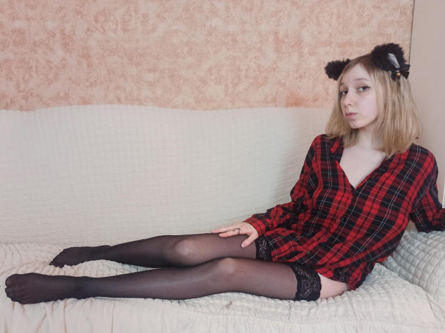 LeahGotty, met blond haar, gekleed in een rood geruit shirt, zwarte kniekousen en zwarte kattenoren, zit op een witte bank, met haar benen uitgestrekt, en ziet er verveeld uit..