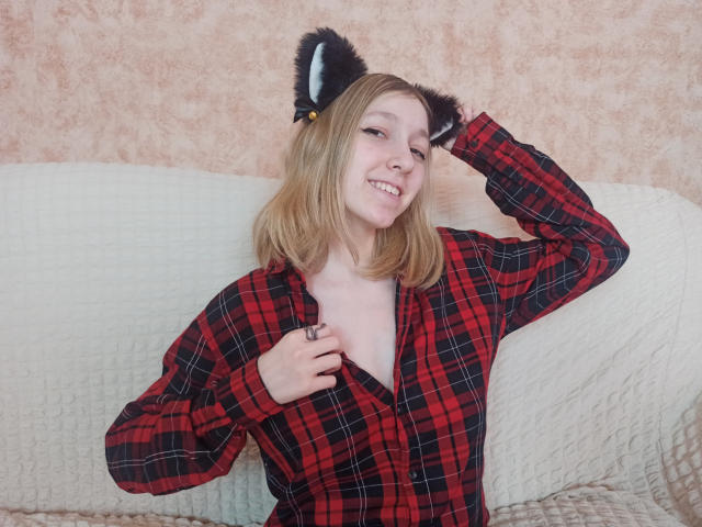 LeahGotty con orejas de gato, sonriendo, vistiendo una camisa de cuadros rojos, mostrando sus pequeños senos, con una mano en el pecho, cabello rubio, sentada en un sofá blanco con textura..