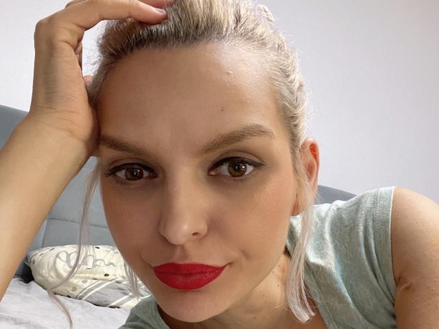 DouceAmelia with blonde hair in a bun, red lipstick, and brown eyes, wearing a grey shirt, hand in hair, looking at camera, slightly smiling.