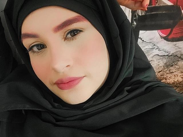 IsabellaStorm, con un hijab negro, lápiz labial rosa y maquillaje de ojos discreto, sostiene una correa negra, tiene una sonrisa suave y cejas rosadas, y su rostro está centrado en la toma..