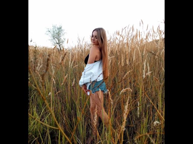 BlondyOneSex com cabelo comprido, top preto com alças, camisa branca aberta, shorts jeans, em pé em um campo alto de trigo dourado, segurando uma lata vermelha, sorrindo olhando por cima do ombro..