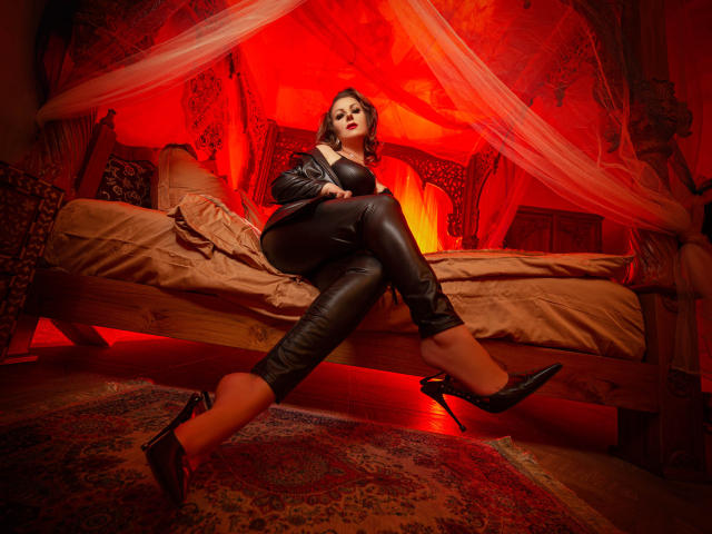 VelvetManttis with big tits, in black leather pants and heels, sitting on a bed, legs crossed, under red lighting, looking seductive, in a red-hued, ornate room.