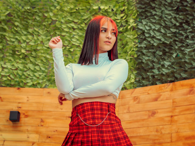 LindaWatsong with shoulder-length black and red hair, white long-sleeve crop top, and red plaid skirt, arms crossed, confident, standing in front of wooden wall.