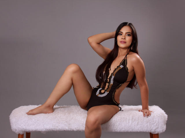 AlexaBlakee, avec ses longs cheveux noirs, est assise sur un banc blanc et moelleux, vêtue d'une combinaison noire avec de profondes découpes latérales qui exposent ses seins et montrent ses fesses..