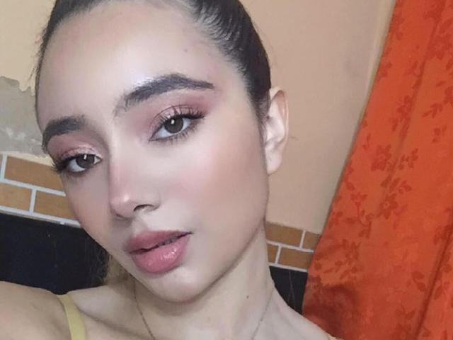 MajoGomez heeft een lichte huid, donkere wenkbrauwen, roze oogschaduw en roze lippen. Ze kijkt naar de camera met een lichte glimlach en draagt een gele top. Haar haar zit in een paardenstaart..