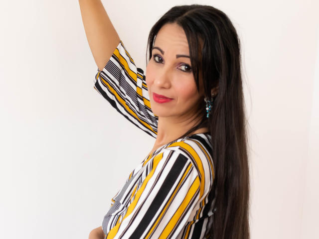 LauraFoxh, com cabelos longos e pretos, blusa listrada amarela, preta e branca, brincos azuis, batom vermelho, sorrindo, braço levantado, mostrando os seios levemente, fundo branco, olhando para a câmera..