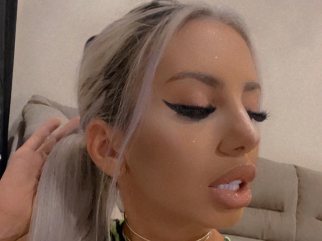 LisaaKiraa has shiny blonde hair in a ponytail, thick black eyeliner, and glossy pink lips. Her skin is tan with subtle sparkles. Her eyes are closed and her lips are slightly parted.