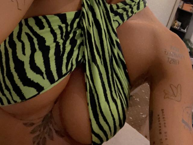 LisaaKiraa's green and black zebra-striped bikini is pulled to the side, revealing her large, round, tan-skinned boobs with a tattoo of flowers on her lower belly.