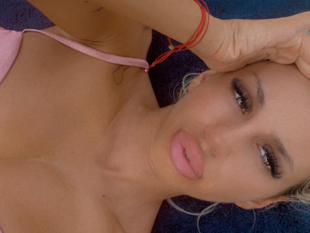 LisaaKiraa tiene labios rosados, ojos marrones y cabello rubio, con una pulsera roja en su muñeca, vistiendo una blusa rosa que deja ver sus senos, y está acostada..