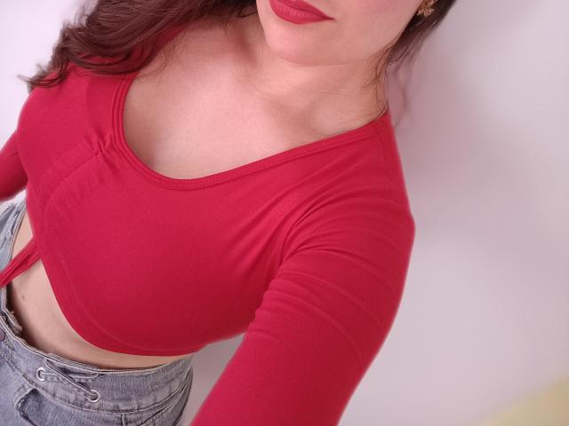 VaaleryStark with red long-sleeve top, showing her tits, in tight fabric, and blue jeans, red lipstick, and wavy brown hair, taking a selfie.