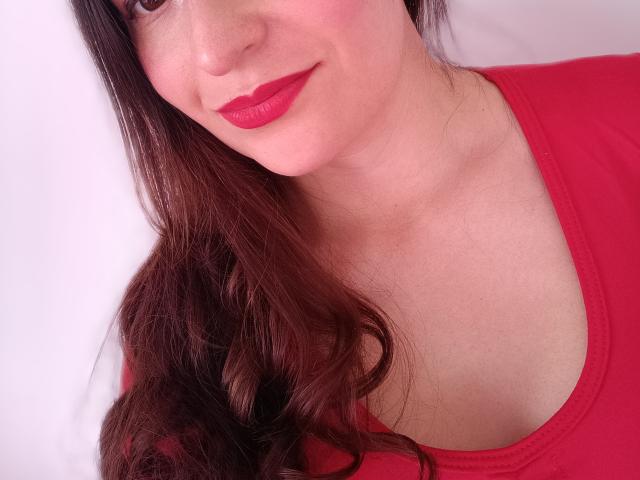 VaaleryStark with red lipstick, wavy brown hair, and red top, showing her tits in a slight upward angle, smiling subtly.