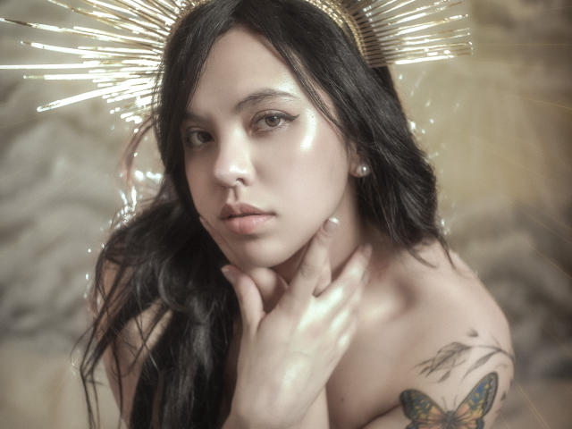 ElyMoon tiene el pelo largo y negro, un rostro brillante y un halo dorado con rayos. Está sin ropa, mostrando un tatuaje de una mariposa en su hombro derecho. Su mano toca su cuello..