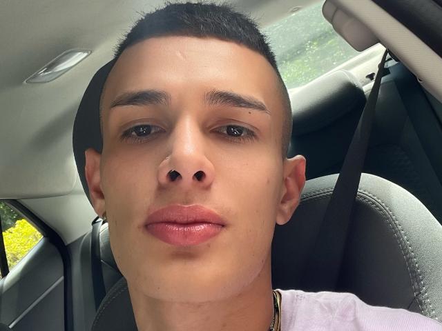 SlimLewis has a cute, tan face with short black hair, brown eyebrows, and glossy pink lips, wearing a white shirt and gold necklace, sitting in a car.