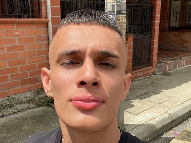 SlimLewis has short brown hair, tan skin, small nose, full pink lips, and a slight smirk. He's wearing a black shirt, standing on a sunny brick sidewalk.