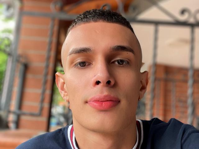 SlimLewis has a clean-shaven face with short brown hair, small earrings, and pink lips. He's wearing a dark blue shirt with white and red trim.