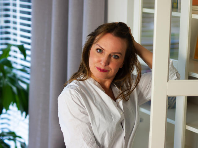 RachelClassy with brown hair, blue eyes, and red lipstick, wearing a white shirt, leaning on a white shelf, hand in hair, smiling, green plant in background, daytime.