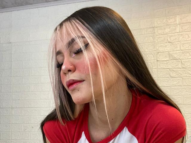Jazmin Wells ha lunghi capelli neri con riflessi rosa, indossa una maglietta raglan rossa e bianca, ha un anello al naso e rossetto rosa, con un leggero sorriso, e guarda di lato..