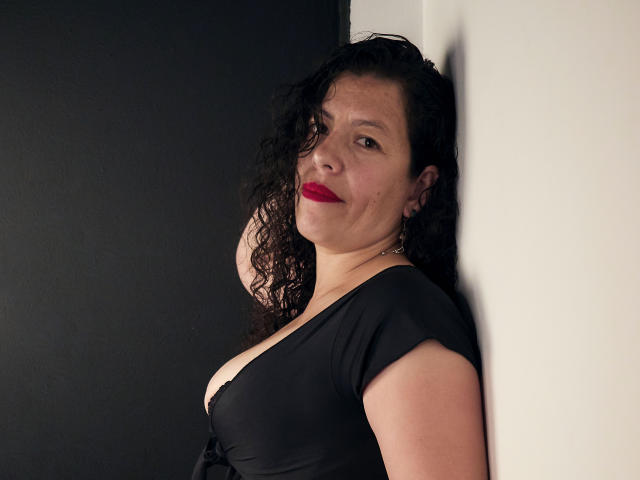 ZalyMature, avec ses cheveux noirs bouclés, son rouge à lèvres rouge et sa robe noire, appuyée contre un mur, montrant sa poitrine dans un haut à décolleté bas, souriant légèrement, avec une expression confiante..