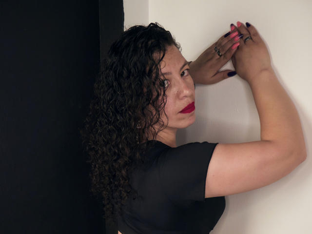 Zaly, con capelli neri ricci, rossetto rosso e maglietta nera, braccia appoggiate contro un muro bianco, che guarda oltre la spalla, con unghie rosa, sfondo nero, espressione seria, mani che toccano il muro..