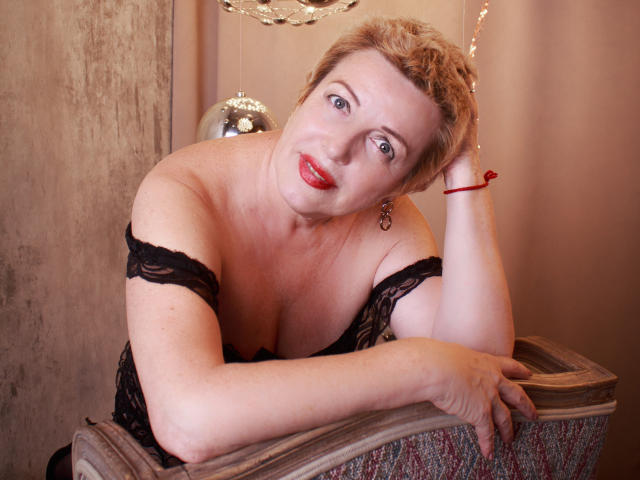SamanthaLala, avec ses cheveux blonds courts, son rouge à lèvres rouge et son haut en dentelle noire, repose sa tête sur une table en bois, montrant sa poitrine généreuse et son bracelet rouge..