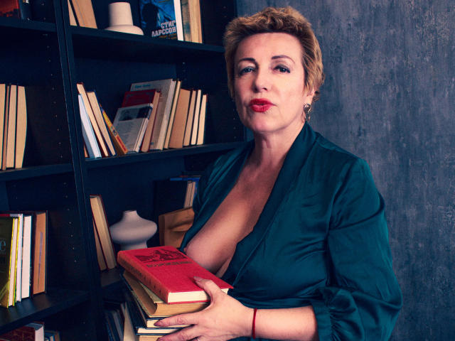 SamanthaLala with short blonde hair, wearing a deep green shirt, exposing her big tits, holding a red book, standing in front of a bookshelf. Her red lips are slightly parted.