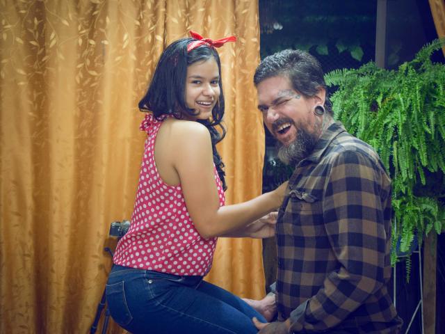 NiaAndDiamond mit dunklem Haar, einem roten Top mit Punkten und Jeans, kniet, lächelt und berührt die Brust eines Mannes mit Bart, der ein kariertes Hemd trägt. Beide lachen, und Nia's Hintern ist sichtbar, in einer gemütlichen Innenraumumgebung..