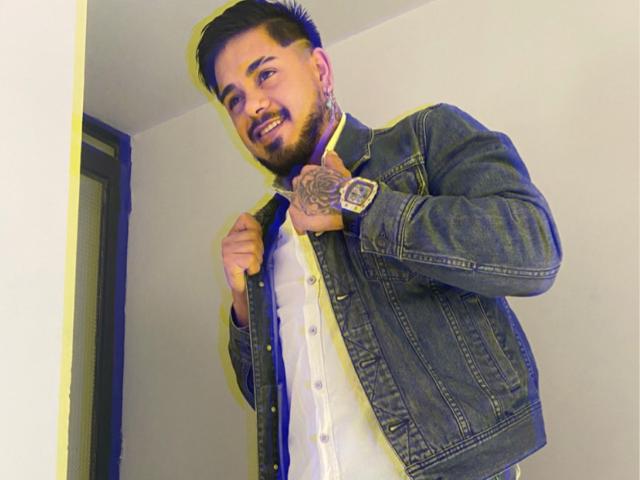 VisiI with short dark hair, beard, and tattoo on right hand, wearing denim jacket over white shirt, smiling, holding jacket collar, black wristwatch, standing in room.