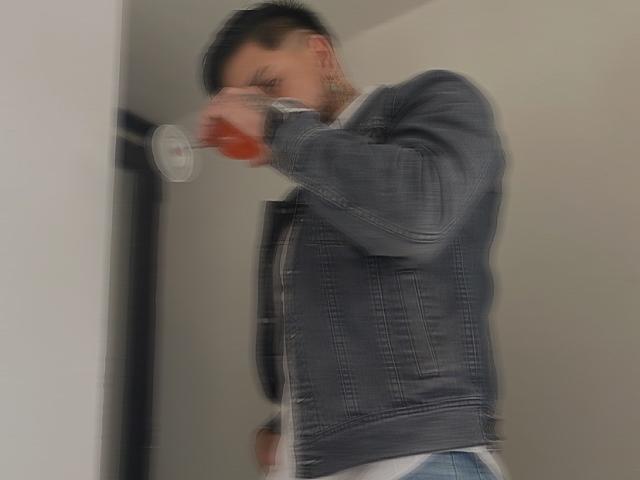 VisiI, com cabelo preto curto, jaqueta jeans e calça jeans azul, bebendo de um copo de vidro transparente com um líquido laranja, olhos parcialmente fechados, em uma foto levemente borrada..