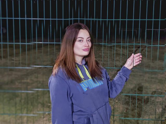 MilanaPrice, mit langen, braunen Haaren, trägt eine blaue Kapuzenjacke und steht vor einem grünen Metallzaun. Sie hält einen schwarzen Stift in ihrer rechten Hand und blickt leicht nach links..