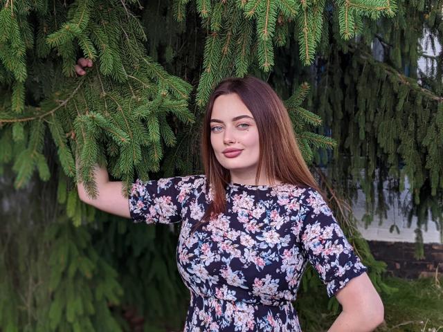MilanaPrice with brown hair, floral dress, and small tits, holding green pine branches with her right hand, smiling softly. She's standing outdoors, surrounded by lush greenery.